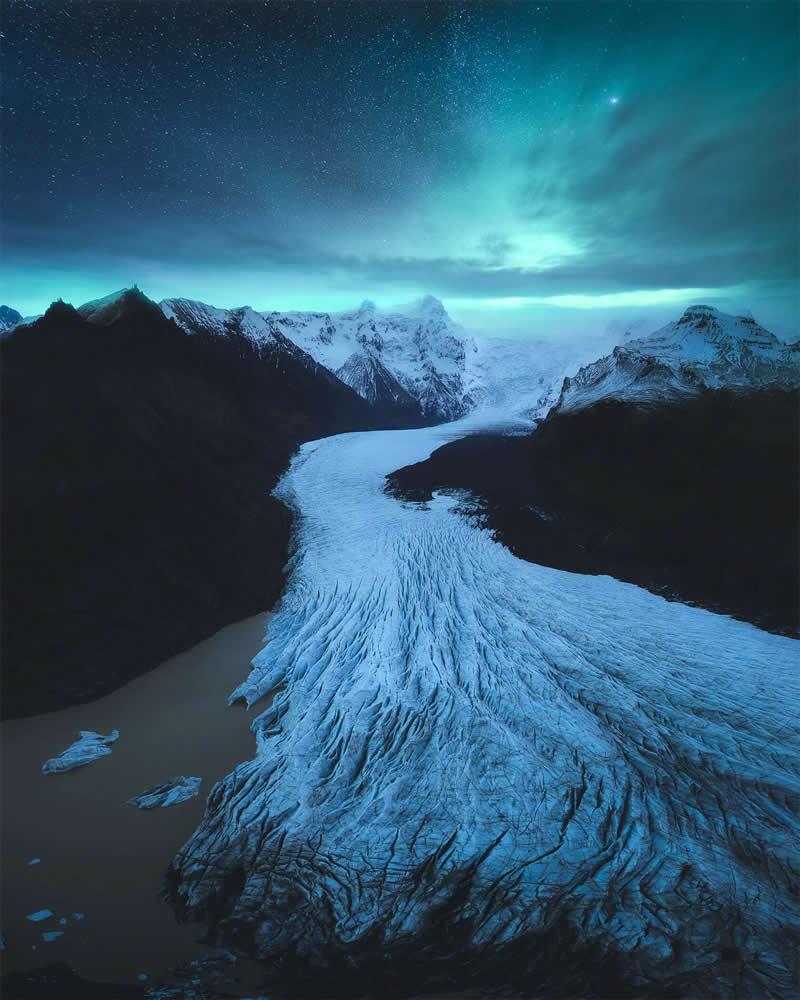 An aerial view of a massive glacier flowing through Iceland's snow-covered mountains beneath the glowing Northern Lights and a star-filled night sky, showcasing a dramatic Arctic landscape.