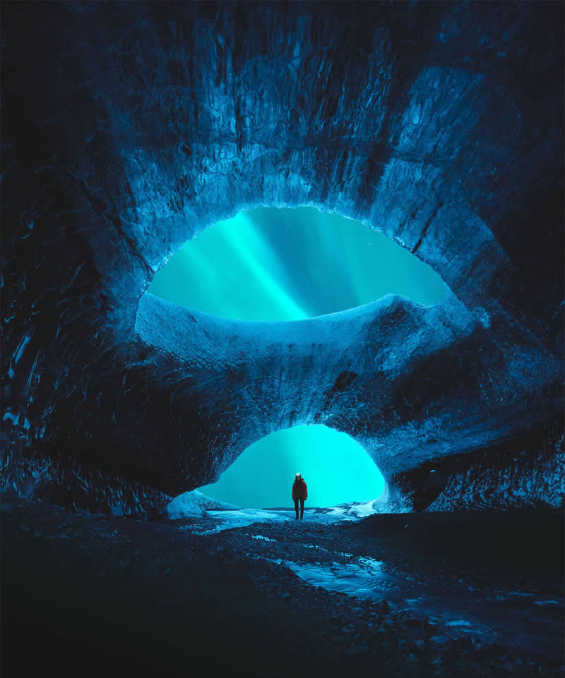 A lone explorer stands inside a vast blue ice cave in Iceland, framed by two glowing openings that reveal the Northern Lights, creating a dramatic and beautifully composed Arctic landscape.
