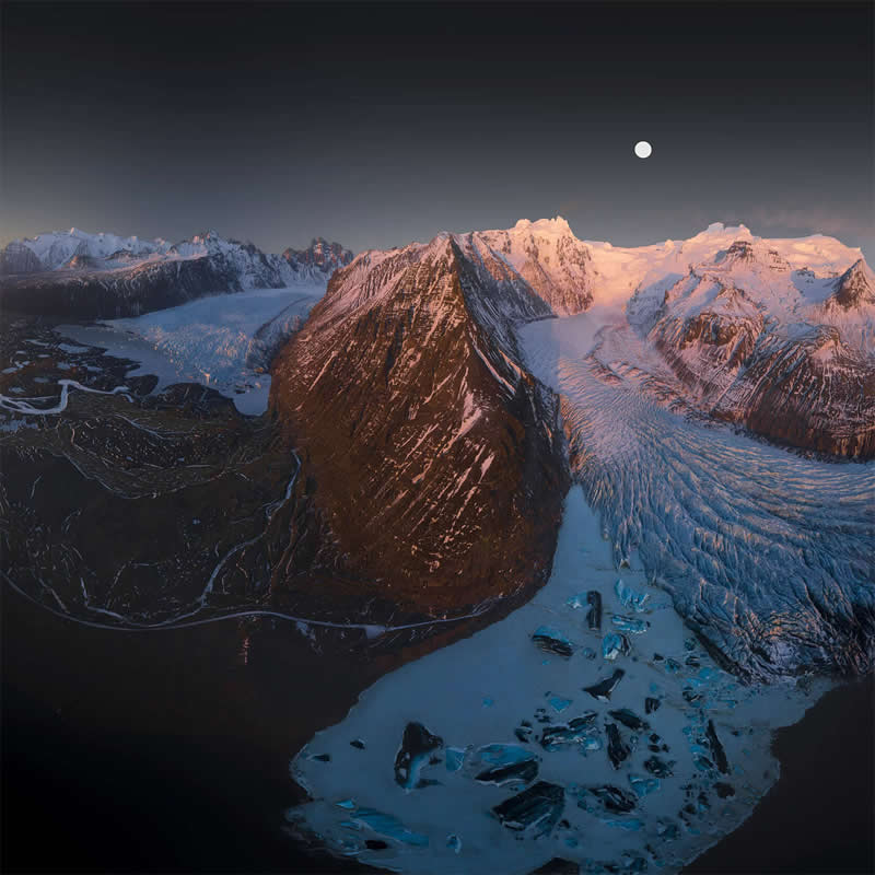 An aerial view of Iceland's snow-covered mountains, sprawling glacier, and icy lagoon beneath a rising moon at twilight, showcasing dramatic landscapes, rugged peaks, and stunning Arctic scenery.