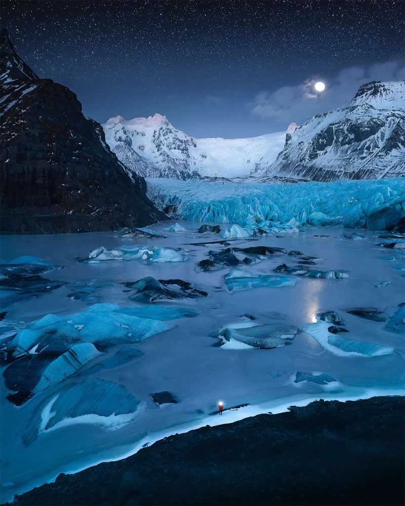 A lone explorer with a headlamp stands beside a frozen glacial lagoon in Iceland beneath a full moon, surrounded by floating blue ice, towering snow-covered mountains, and a star-filled night sky.