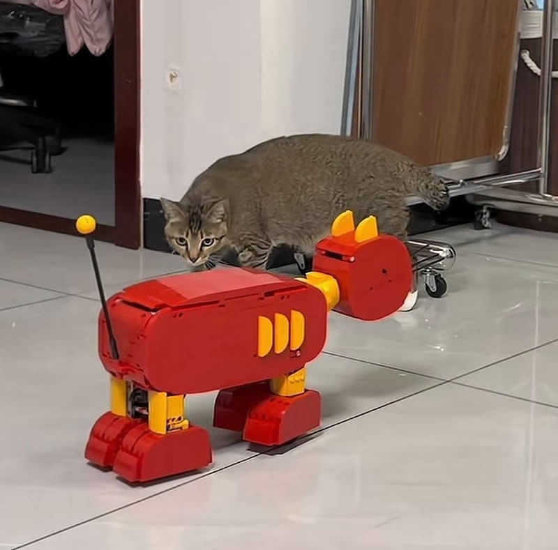 A curious tabby cat crouches low on a tiled floor, carefully stalking a bright red robotic toy. With wide eyes and a cautious pose, the adorable feline looks ready to investigate its mysterious new "robotic rival.