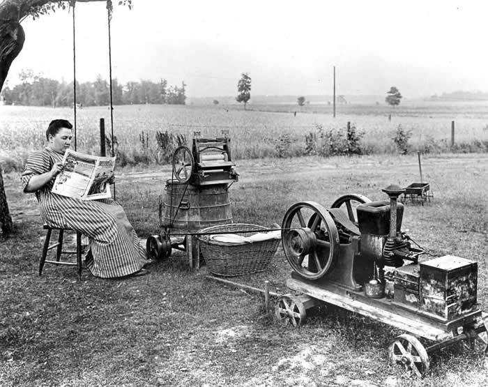 Woman reads newspaper while an early gas powered washing machine works, West Lafayette, Indiana, 1914.