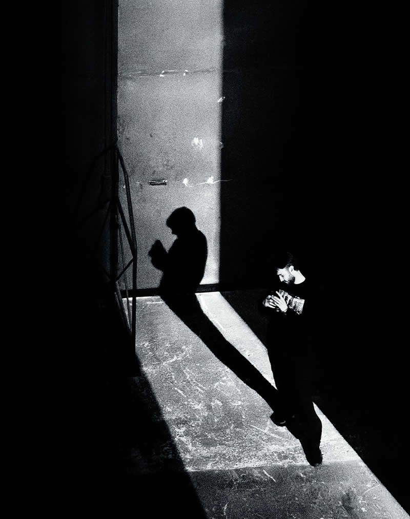 Black-and-white street photograph of a person standing in a bright shaft of light surrounded by deep shadows. The subject&rsquo;s elongated shadow stretches across the illuminated floor, creating dramatic contrast, strong geometry, and a perfectly timed urban scene.