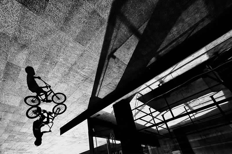 Black-and-white street photograph of a cyclist and their reflection captured on a reflective surface beneath an urban structure. Dramatic shadows, geometric lines, and an unusual perspective create a striking composition emphasizing timing, symmetry, and urban design.