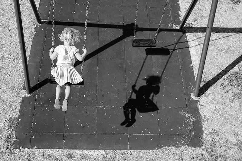 Black-and-white photograph of a young girl on a swing viewed from above. Her shadow appears on the playground surface beside an empty swing, creating a striking visual relationship through light, timing, and composition.
