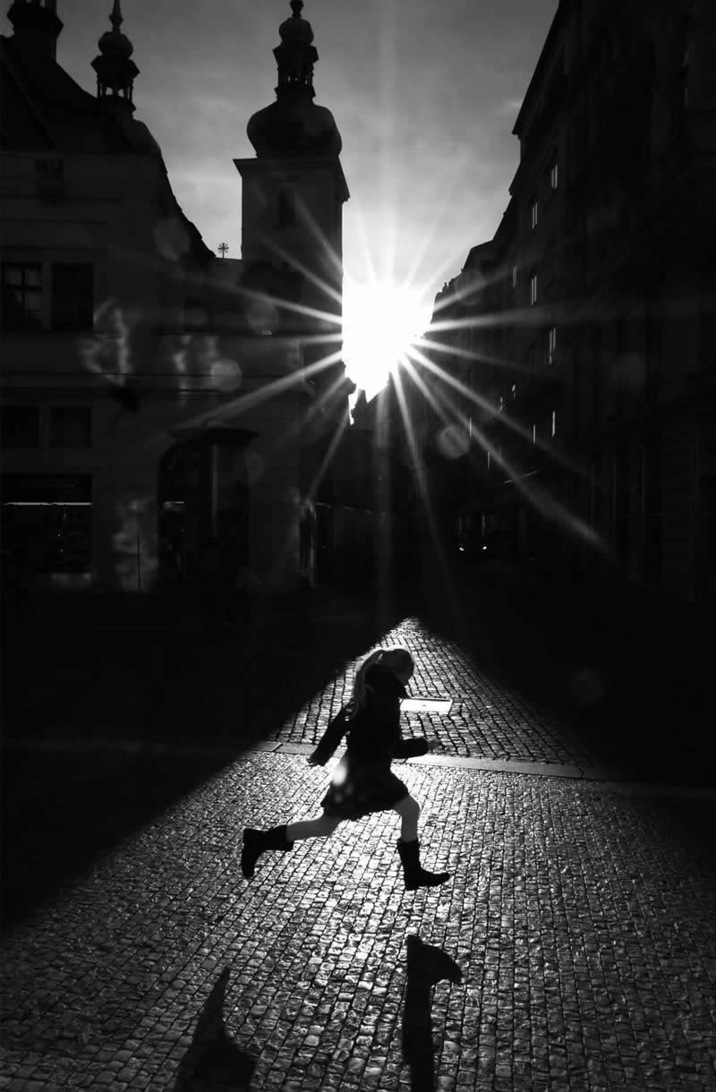 Black-and-white street photograph of a person running across a cobblestone square as sunlight bursts between historic buildings. Strong silhouettes, starburst light rays, long shadows, and precise timing create a dramatic and visually striking urban scene.