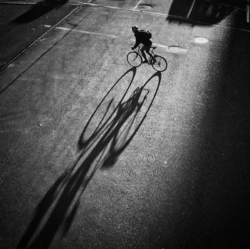 Black-and-white street photograph of a cyclist riding across a sunlit road, casting a long elongated shadow. Viewed from above, the image emphasizes dramatic light, strong contrast, geometric shapes, and a perfectly timed urban moment.
