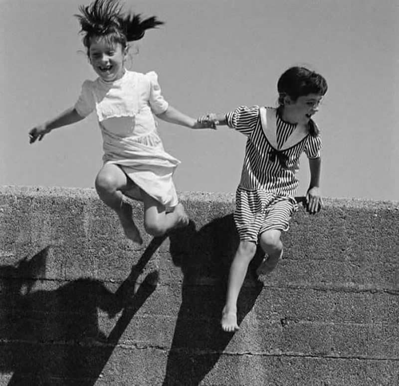Black-and-white photograph of two young girls holding hands while jumping from a stone wall. One child is captured mid-air with hair flying upward, while strong shadows below add depth, movement, and dramatic visual impact.