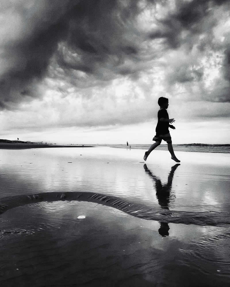 Black-and-white photograph of a child running across a wet beach beneath dramatic storm clouds. Reflections shimmer on the water-covered sand, while the child&rsquo;s silhouette and mirrored image create a dynamic composition filled with movement and atmosphere.