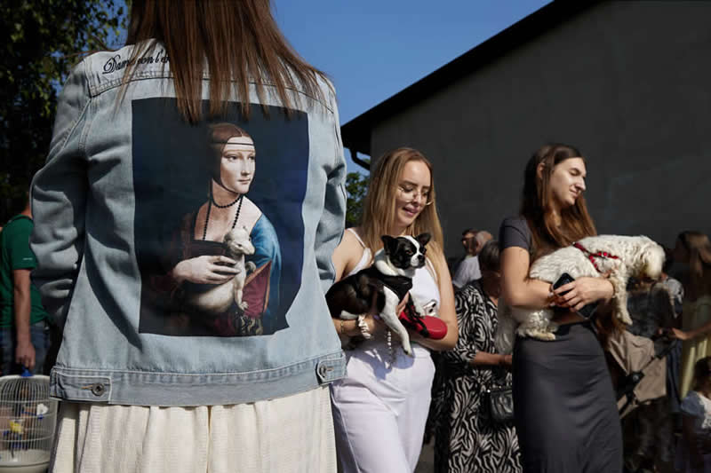 Color street photograph showing a woman wearing a denim jacket printed with a classical portrait holding an animal, while nearby women hold small dogs. Clever timing and framing create a visual connection between the artwork and real-life scene.