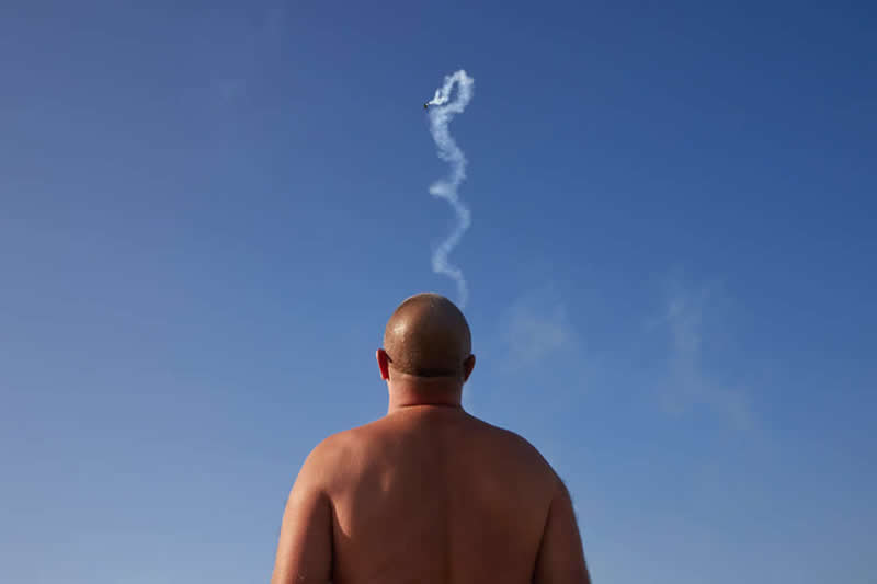 Minimalist photograph of a bald man viewed from behind against a clear blue sky, with a winding smoke trail from an aircraft perfectly aligned above his head, creating the illusion of a thought or plume rising upward.