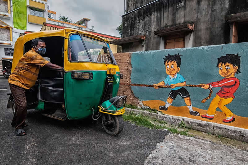 Street photograph of an auto-rickshaw parked beside a colorful wall mural of two children pulling a rope, captured from an angle that makes it appear as though the painted children are pulling the vehicle.