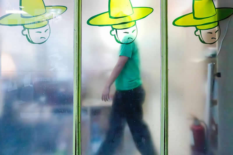 Street photograph of a person walking behind frosted glass panels, perfectly aligned with a cartoon figure wearing a large green hat, creating the illusion that the illustrated hat belongs to the passerby.