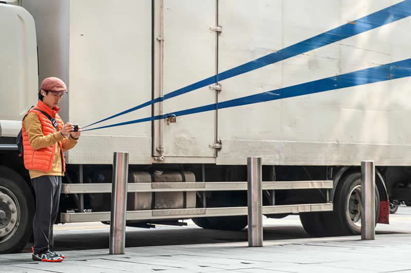 Street photograph of a man holding a camera beside a parked truck, with blue stripes on the truck perfectly aligned to appear as though they extend directly from the camera lens.