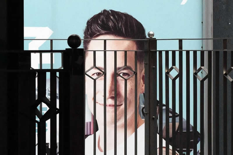 Street photograph of a large billboard portrait partially obscured by a decorative metal fence, with the fence bars aligning across the subject&rsquo;s face to create a striking geometric composition.