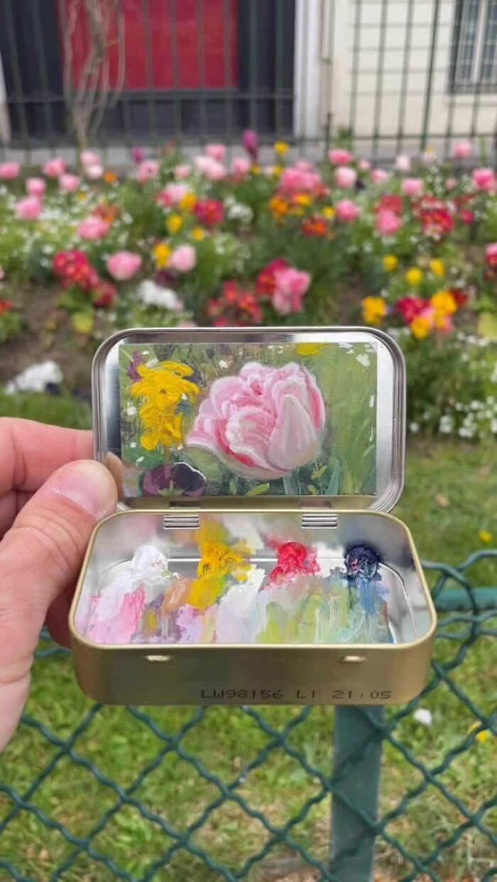 These 40 Tiny Altoid Tin Paintings by Remington Robinson Are Pure Pocket-Sized Magic 45 A hand holds an open Altoid tin in front of a flower garden, revealing a miniature floral painting by Remington Robinson inside the lid, featuring a soft pink blossom and yellow flowers, with a tiny pastel paint palette below.