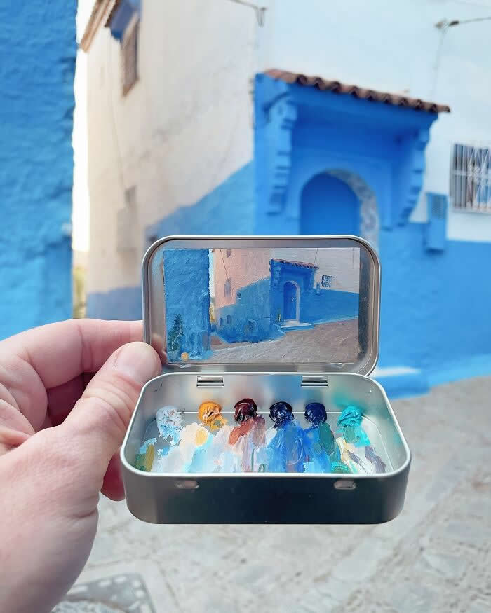 These 40 Tiny Altoid Tin Paintings by Remington Robinson Are Pure Pocket-Sized Magic 46 A hand holds an open Altoid tin in front of a blue-painted street scene, showing a miniature architectural painting by Remington Robinson inside the lid, with a small palette of blue, white, and earth-toned paints arranged in the bottom half.