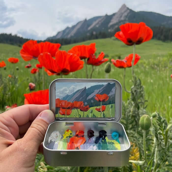These 40 Tiny Altoid Tin Paintings by Remington Robinson Are Pure Pocket-Sized Magic 47 A hand holds an open Altoid tin in a mountain meadow filled with bright red poppies, revealing a miniature landscape painting by Remington Robinson inside the lid, with a tiny paint palette arranged in the bottom half.