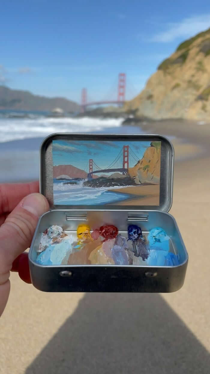 These 40 Tiny Altoid Tin Paintings by Remington Robinson Are Pure Pocket-Sized Magic 49 A hand holds an open Altoid tin on a beach near the Golden Gate Bridge, showing a miniature coastal painting by Remington Robinson inside the lid, with a tiny paint palette of blues, sandy neutrals, and warm reds in the bottom half.