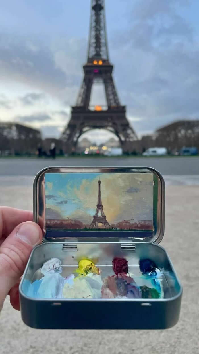 These 40 Tiny Altoid Tin Paintings by Remington Robinson Are Pure Pocket-Sized Magic 51 A hand holds an open Altoid tin in front of the Eiffel Tower at dusk, revealing a miniature painting by Remington Robinson of the landmark under a soft evening sky, with a tiny paint palette arranged in the bottom half.