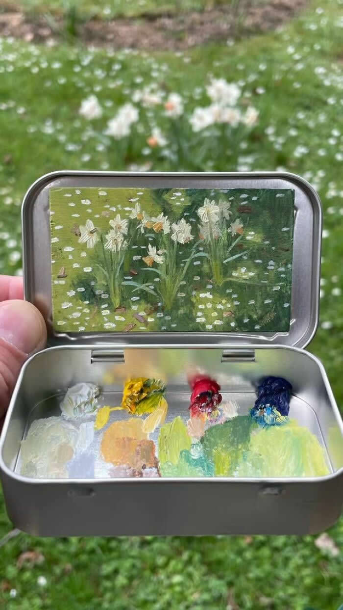 These 40 Tiny Altoid Tin Paintings by Remington Robinson Are Pure Pocket-Sized Magic 52 A hand holds an open Altoid tin in front of a grassy patch dotted with white flowers, revealing a miniature painting by Remington Robinson of blooming daffodils and scattered petals, with a tiny paint palette of greens, whites, yellow, red, and blue in the bottom half.