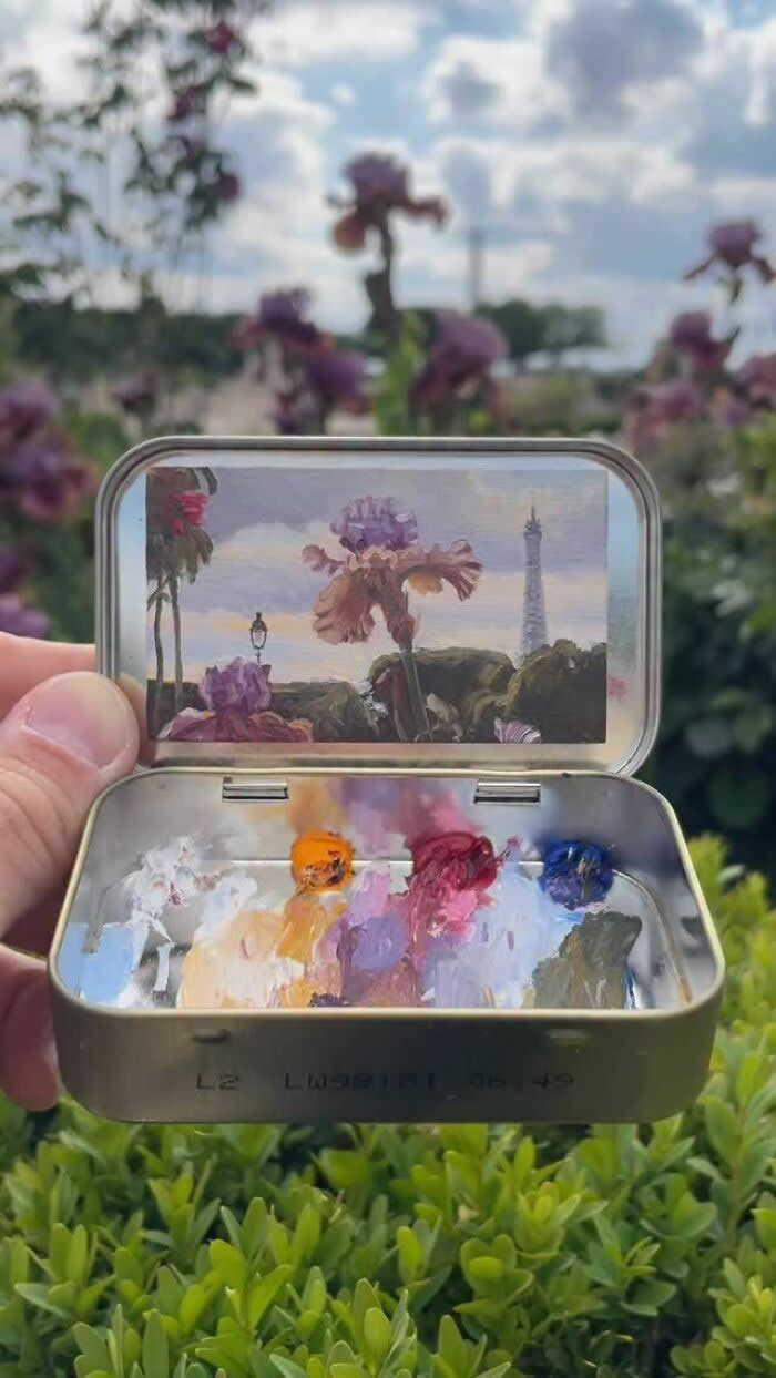 These 40 Tiny Altoid Tin Paintings by Remington Robinson Are Pure Pocket-Sized Magic 53 A hand holds an open Altoid tin in a flower garden, revealing a miniature painting by Remington Robinson of purple blooms with the Eiffel Tower rising in the background, while a small pastel-toned paint palette sits in the bottom half.