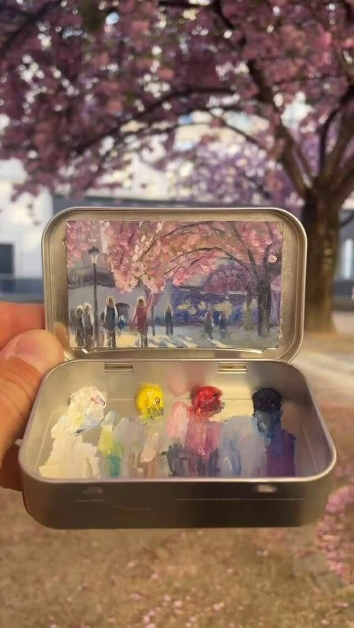 These 40 Tiny Altoid Tin Paintings by Remington Robinson Are Pure Pocket-Sized Magic 54 A hand holds an open Altoid tin beneath blooming pink cherry trees, revealing a miniature painting by Remington Robinson of people walking under a canopy of blossoms, with a tiny pastel paint palette arranged in the bottom half.