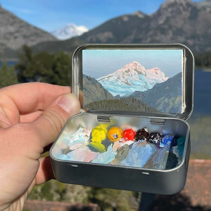 These 40 Tiny Altoid Tin Paintings by Remington Robinson Are Pure Pocket-Sized Magic 56 A hand holds an open Altoid tin in front of a mountain lake view, revealing a miniature painting by Remington Robinson of a snow-capped peak glowing in soft light, with a tiny paint palette of blues, whites, yellows, and warm earth tones in the bottom half.