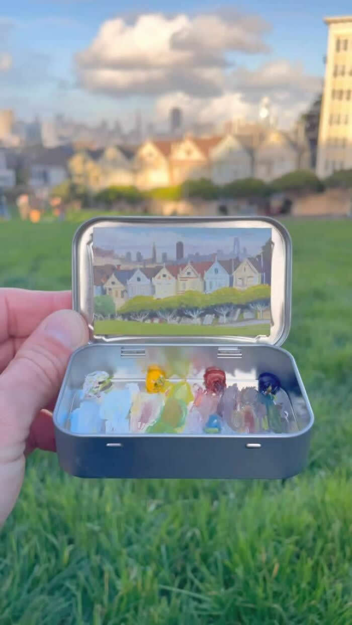 These 40 Tiny Altoid Tin Paintings by Remington Robinson Are Pure Pocket-Sized Magic 57 A hand holds an open Altoid tin in a grassy field, revealing a miniature painting by Remington Robinson of a row of pale houses and trees under a cloudy sky, with a tiny pastel-toned paint palette arranged in the bottom half.