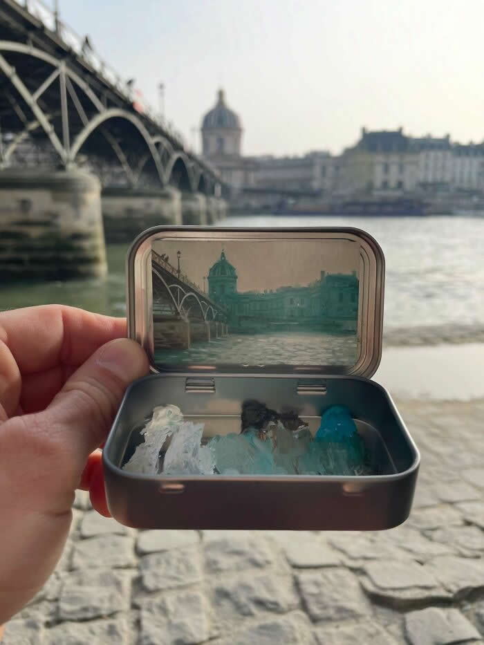 These 40 Tiny Altoid Tin Paintings by Remington Robinson Are Pure Pocket-Sized Magic 60 A hand holds an open Altoid tin beside a riverside in Paris, revealing a miniature painting by Remington Robinson of a bridge and domed building reflected on the Seine, with a tiny cool-toned paint palette arranged in the bottom half.