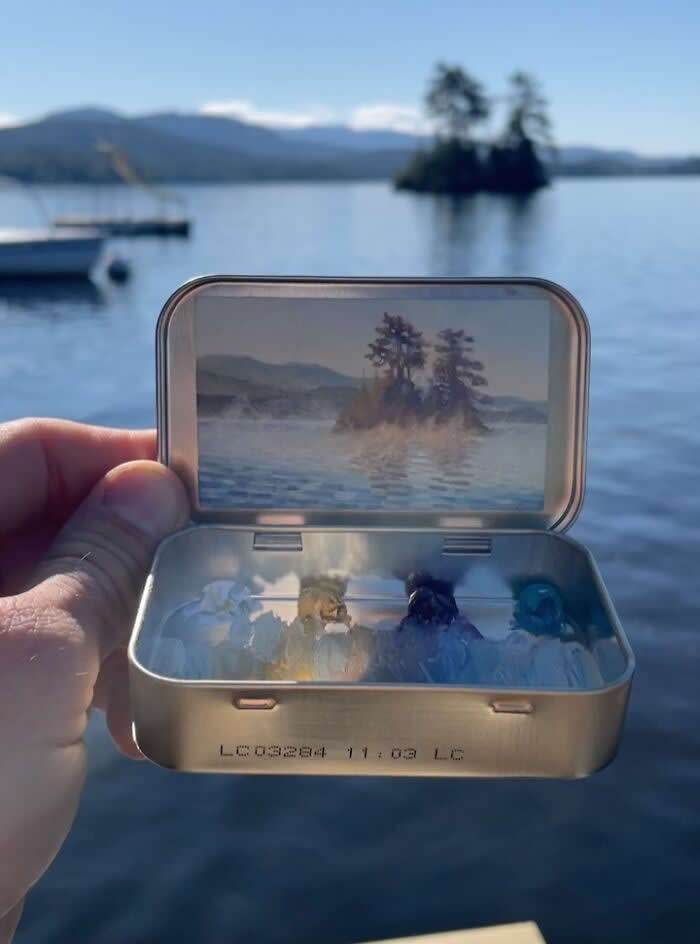 These 40 Tiny Altoid Tin Paintings by Remington Robinson Are Pure Pocket-Sized Magic 62 A hand holds an open Altoid tin beside a calm blue lake, revealing a miniature painting by Remington Robinson of a small tree-covered island rising through soft morning mist, with a tiny cool-toned paint palette arranged in the bottom half.