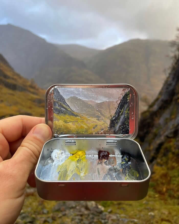 These 40 Tiny Altoid Tin Paintings by Remington Robinson Are Pure Pocket-Sized Magic 63 A hand holds an open Altoid tin in a misty mountain valley, revealing a miniature landscape painting by Remington Robinson of green slopes and a winding glen after rain, with a tiny paint palette of white, yellow, earthy browns, and gray in the bottom half.