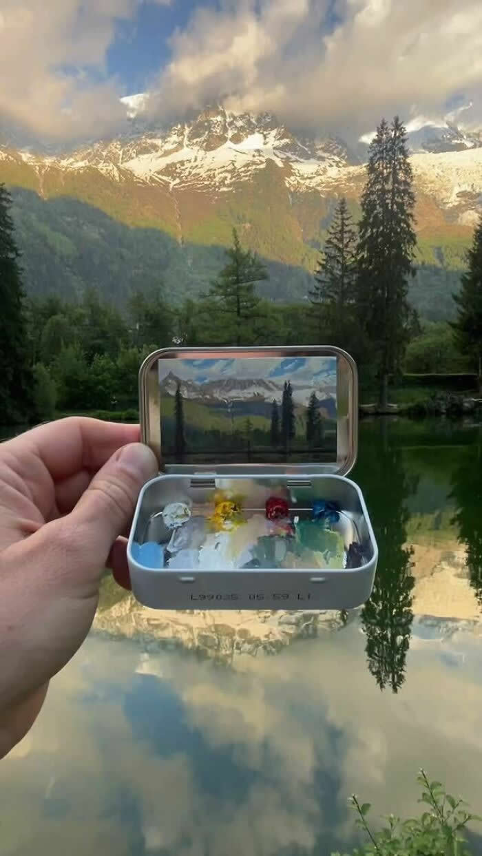 These 40 Tiny Altoid Tin Paintings by Remington Robinson Are Pure Pocket-Sized Magic 64 A hand holds an open Altoid tin in front of a calm alpine lake, revealing a miniature landscape painting by Remington Robinson of snow-capped mountains, tall trees, and their reflections in the water, with a tiny paint palette arranged in the bottom half.