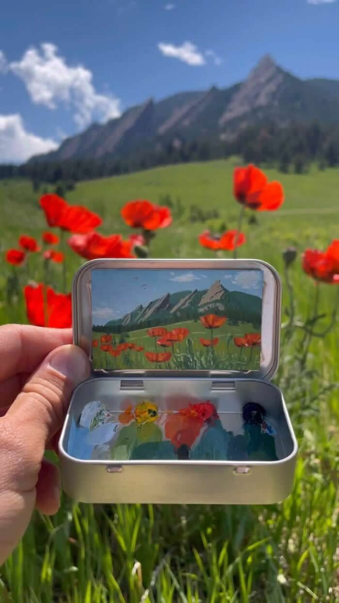 These 40 Tiny Altoid Tin Paintings by Remington Robinson Are Pure Pocket-Sized Magic 65 A hand holds an open Altoid tin in a mountain meadow filled with vivid red poppies, revealing a miniature landscape painting by Remington Robinson of the same wildflower field with jagged peaks in the distance, with a tiny paint palette arranged in the bottom half.