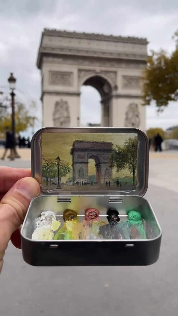 These 40 Tiny Altoid Tin Paintings by Remington Robinson Are Pure Pocket-Sized Magic 67 A hand holds an open Altoid tin in front of the Arc de Triomphe, revealing a miniature painting by Remington Robinson of the Paris monument and nearby trees under a muted sky, with a tiny paint palette of white, olive, pink, black, and green in the bottom half.