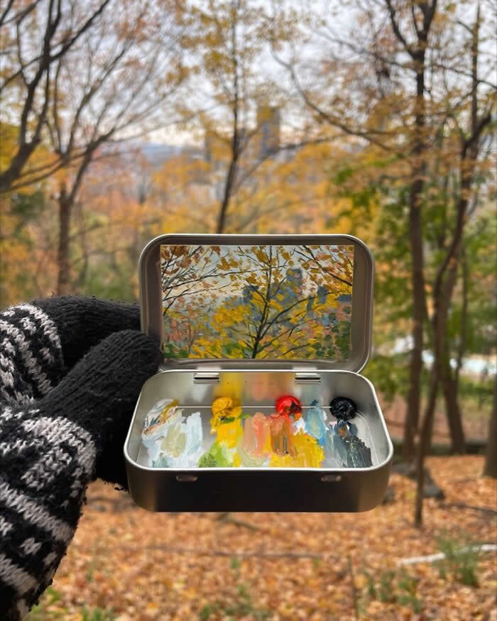 These 40 Tiny Altoid Tin Paintings by Remington Robinson Are Pure Pocket-Sized Magic 68 A gloved hand holds an open Altoid tin in a quiet autumn woodland, revealing a miniature painting by Remington Robinson of golden leaves and slender tree branches, with a tiny paint palette of whites, yellows, reds, blues, and black in the bottom half.