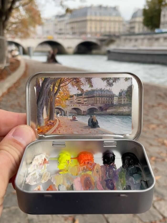 These 40 Tiny Altoid Tin Paintings by Remington Robinson Are Pure Pocket-Sized Magic 69 A hand holds an open Altoid tin beside the Seine in Paris, revealing a miniature painting by Remington Robinson of a riverside walkway lined with autumn trees and a stone bridge, with a tiny paint palette of white, yellow, orange, purple, and black in the bottom half.