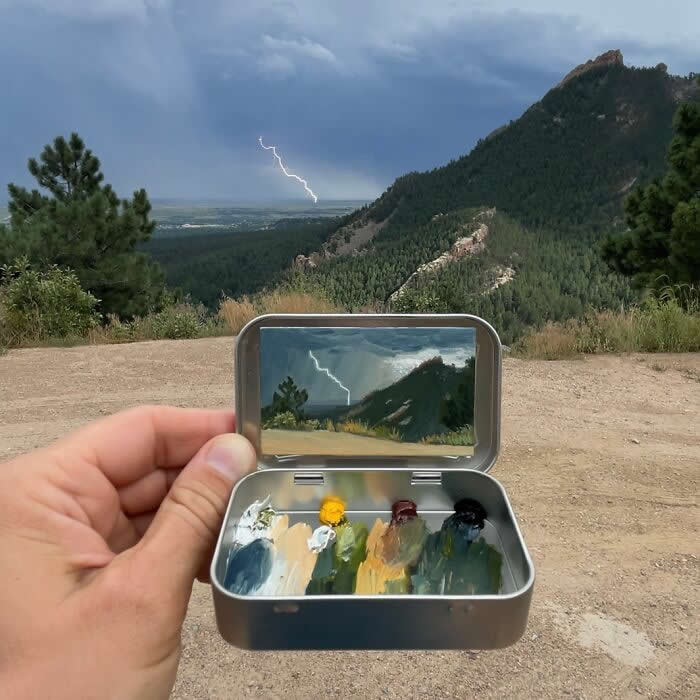 These 40 Tiny Altoid Tin Paintings by Remington Robinson Are Pure Pocket-Sized Magic 70 A hand holds an open Altoid tin at a mountain overlook during a storm, revealing a miniature landscape painting by Remington Robinson of a lightning strike cutting across a dark sky above forested hills, with a tiny paint palette of blues, greens, yellow, and earthy tones in the bottom half.