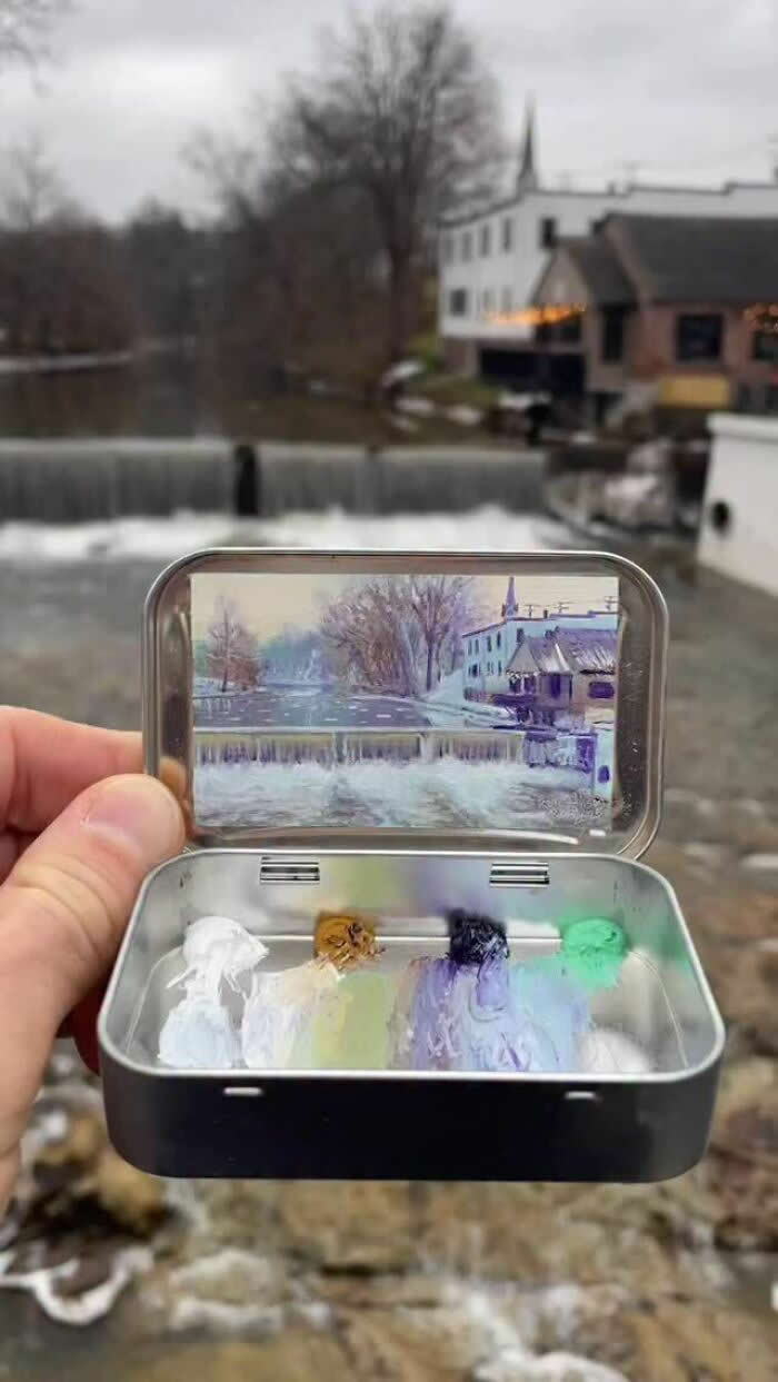 These 40 Tiny Altoid Tin Paintings by Remington Robinson Are Pure Pocket-Sized Magic 72 A hand holds an open Altoid tin beside a small riverside waterfall on a gray winter day, revealing a miniature painting by Remington Robinson of snow-dusted buildings, bare trees, and cascading water, with a tiny paint palette of white, ochre, violet, and mint green in the bottom half.