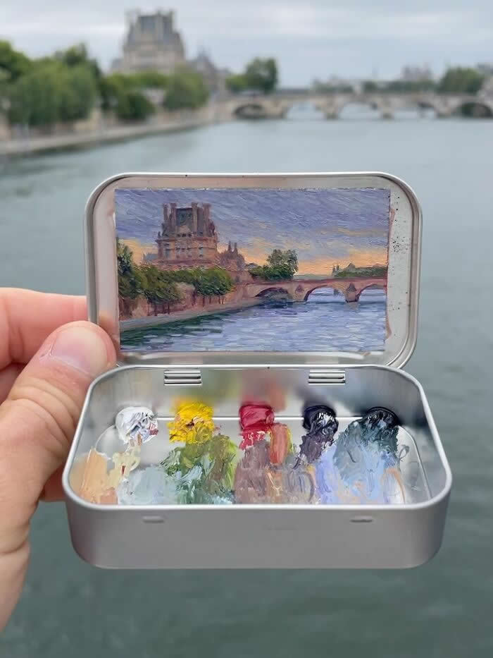 These 40 Tiny Altoid Tin Paintings by Remington Robinson Are Pure Pocket-Sized Magic 73 A hand holds an open Altoid tin beside the Seine in Paris, revealing a miniature painting by Remington Robinson of Notre-Dame and a stone bridge glowing under a soft evening sky, with a tiny paint palette of white, yellow, crimson, and cool blue-gray tones in the bottom half.