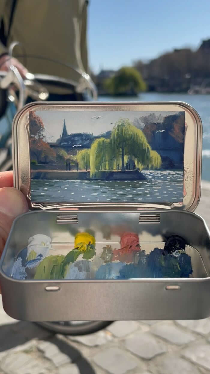 These 40 Tiny Altoid Tin Paintings by Remington Robinson Are Pure Pocket-Sized Magic 74 A hand holds an open Altoid tin beside a sunlit river, revealing a miniature painting by Remington Robinson of a weeping willow reflected on shimmering water, with a tiny paint palette of white, yellow, green, pink, blue, and black in the bottom half.