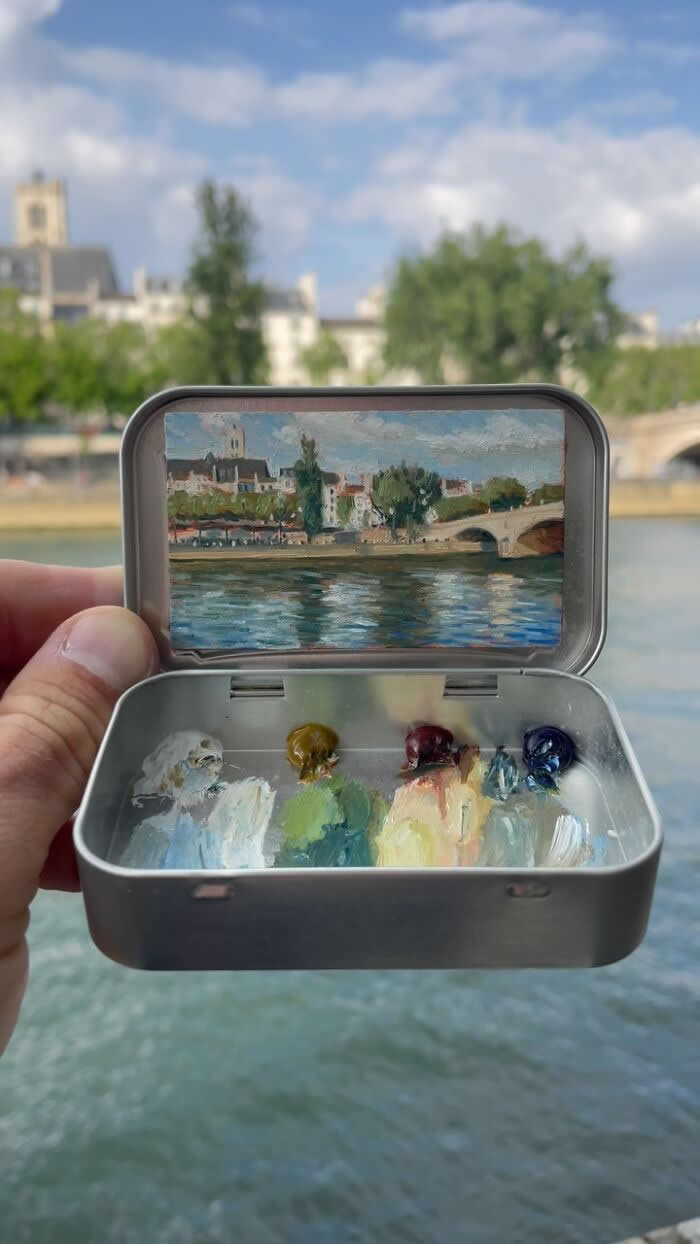 These 40 Tiny Altoid Tin Paintings by Remington Robinson Are Pure Pocket-Sized Magic 75 A hand holds an open Altoid tin beside a calm river, revealing a miniature painting by Remington Robinson of a sunlit riverside town with a stone bridge and shimmering reflections on the water, with a tiny paint palette of white, olive, burgundy, cream, and blue in the bottom half.