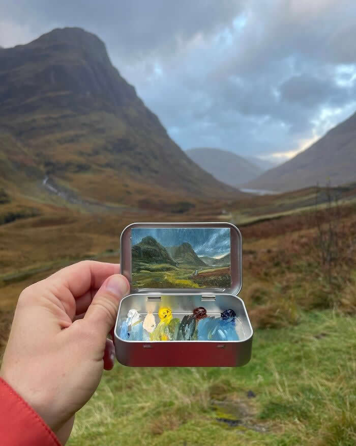 These 40 Tiny Altoid Tin Paintings by Remington Robinson Are Pure Pocket-Sized Magic 76 A hand holds an open Altoid tin in a dramatic mountain valley, revealing a miniature landscape painting by Remington Robinson of rolling green slopes, winding water, and dark peaks beneath a moody sky, with a tiny paint palette of white, yellow, brown, blue, and green in the bottom half.