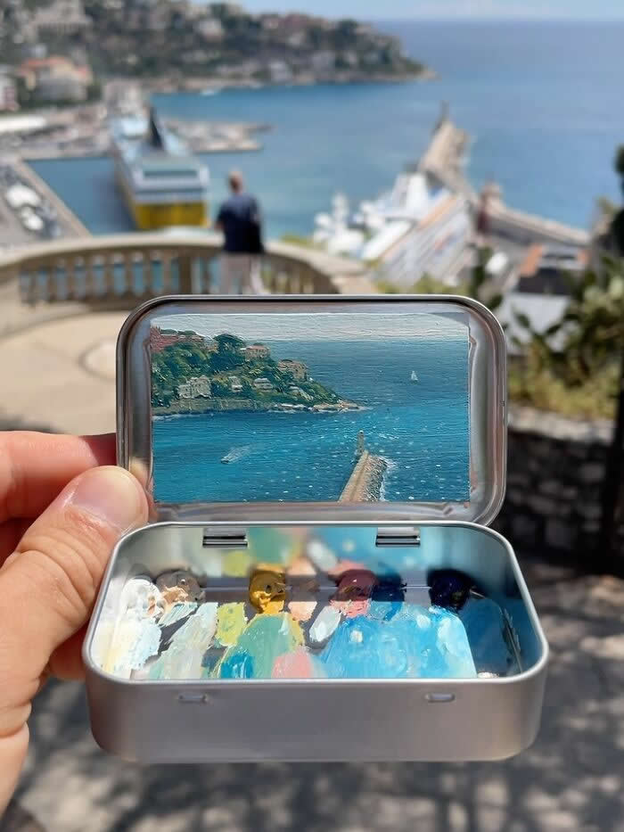 These 40 Tiny Altoid Tin Paintings by Remington Robinson Are Pure Pocket-Sized Magic 79 A hand holds an open Altoid tin overlooking a bright coastal harbor, revealing a miniature painting by Remington Robinson of turquoise water, a long pier, and a hillside dotted with seaside buildings, with a tiny paint palette of white, tan, gold, pink, and deep blue in the bottom half.