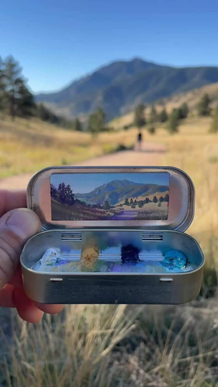 These 40 Tiny Altoid Tin Paintings by Remington Robinson Are Pure Pocket-Sized Magic 80 A hand holds an open Altoid tin in a sunlit mountain valley, revealing a miniature landscape painting by Remington Robinson of a winding path, golden grass, and distant blue peaks, with a tiny paint palette of pale yellow, violet, deep blue, and turquoise in the bottom half.
