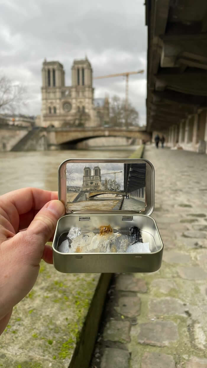 These 40 Tiny Altoid Tin Paintings by Remington Robinson Are Pure Pocket-Sized Magic 81 A hand holds an open Altoid tin along the Seine in Paris, revealing a miniature painting by Remington Robinson of Notre-Dame, a stone bridge, and the riverside walkway under an overcast sky, with a tiny neutral-toned paint palette in the bottom half.