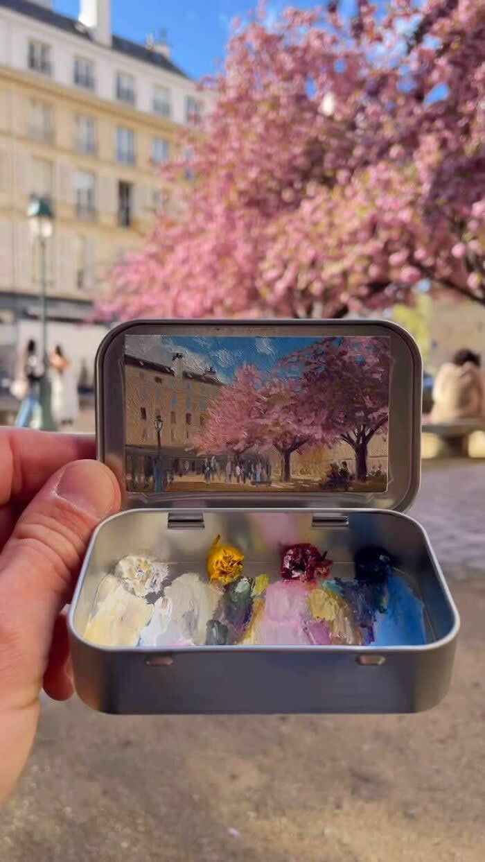 These 40 Tiny Altoid Tin Paintings by Remington Robinson Are Pure Pocket-Sized Magic 82 A hand holds an open Altoid tin in a sunlit square lined with pink cherry blossoms, revealing a miniature painting by Remington Robinson of pastel buildings, flowering trees, and people strolling beneath the blooms, with a tiny paint palette of white, yellow, pink, burgundy, and blue in the bottom half.