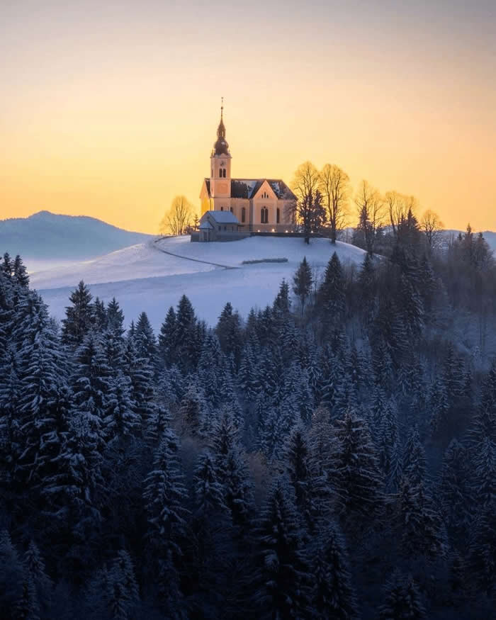 Beauty of the Earth: 33 Epic Landscape Views That Prove Nature Is the Greatest Artist 36 A church stands on a snowy hill at sunrise above a dense dark pine forest, with soft golden sky and distant mountain silhouettes in the background.