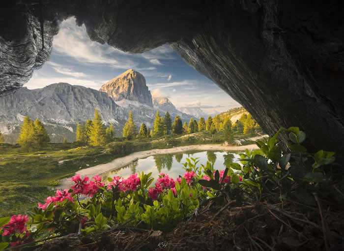 Beauty of the Earth: 33 Epic Landscape Views That Prove Nature Is the Greatest Artist 40 A mountain peak and small reflective pond are seen through the opening of a dark cave, with bright pink flowers and green plants in the foreground under soft daylight.
