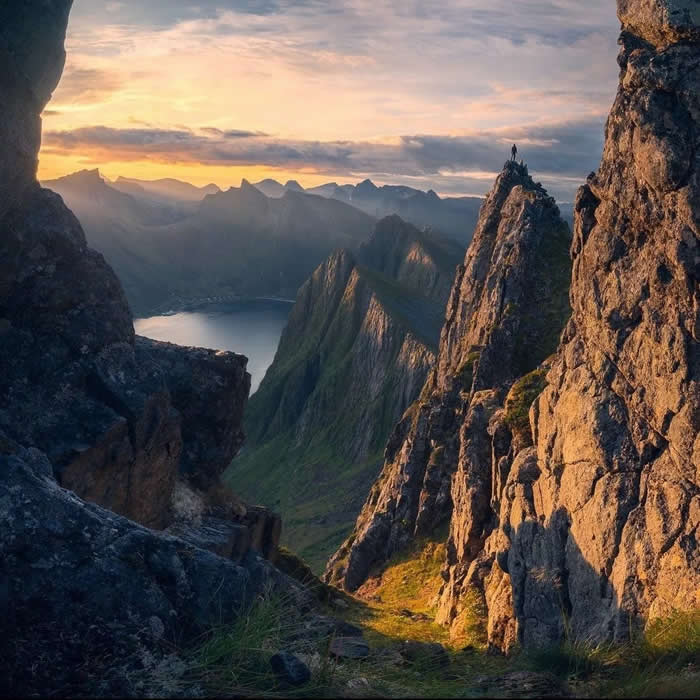 Beauty of the Earth: 33 Epic Landscape Views That Prove Nature Is the Greatest Artist 41 A dramatic mountain landscape at sunset seen between rocky cliffs, with a lone person standing on a sharp peak above a fjord and distant mountain ridges.